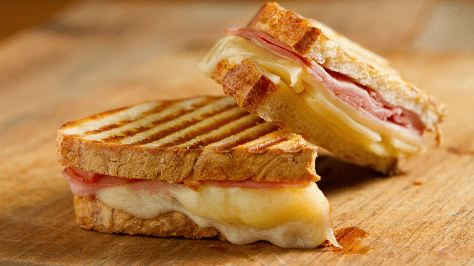 Grilled ham and cheese sandwich cut in half and on a wooden cutting board. The bread on the sandwich has grill marks from a panini press.