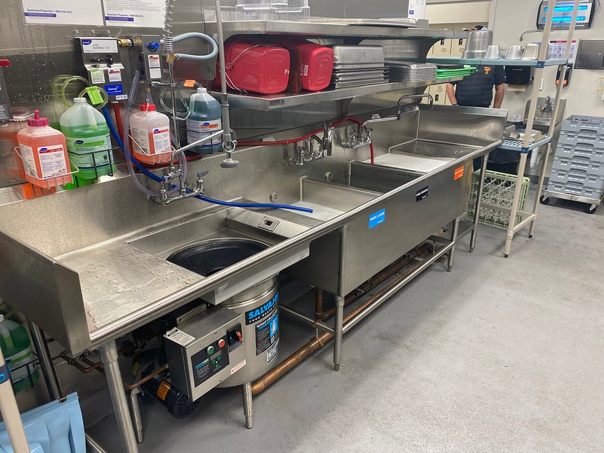 Titan Stainless sinks in back of commercial kitchen. Jugs of cleaning and sanitizing solutions hang behind on wall and red buckets on shelves above.